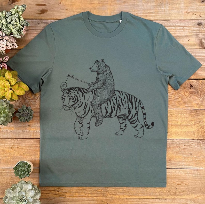 May include: A green t-shirt with a black and white illustration of a bear riding a tiger. The bear is holding a stick with a circle on the end.