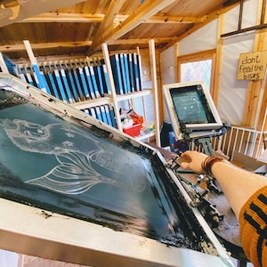 May include: A close-up of a screen printing process with a white mermaid design on a black screen. The screen is being held by a person wearing a brown bracelet and a yellow and black striped sweater. A sign in the background reads "Don't feed the bears."