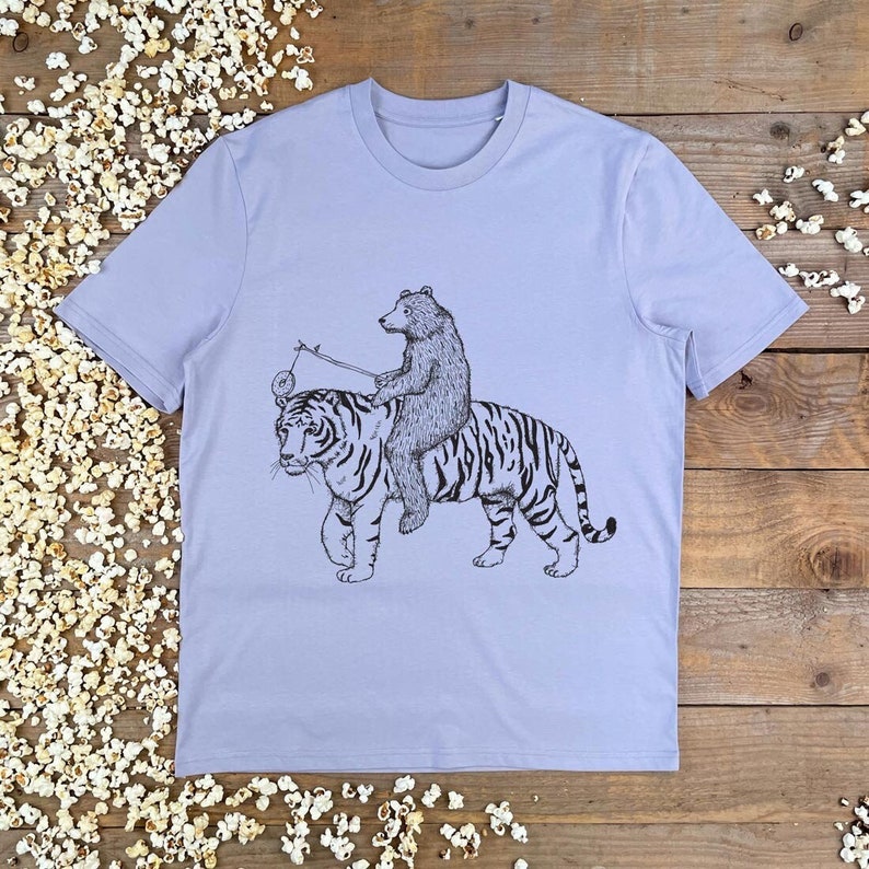 May include: A lavender t-shirt with a black and white illustration of a bear riding a tiger. The bear is holding a stick with a ring on the end.