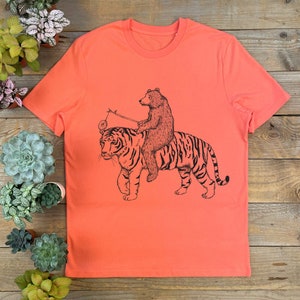 May include: A coral orange t-shirt with a black and white illustration of a bear riding a tiger. The bear is holding a stick with a ball on the end.