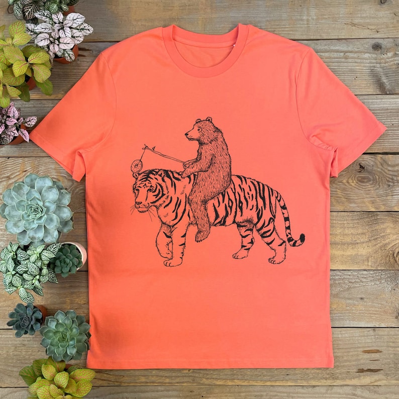May include: A coral orange t-shirt with a black and white illustration of a bear riding a tiger. The bear is holding a stick with a ball on the end.