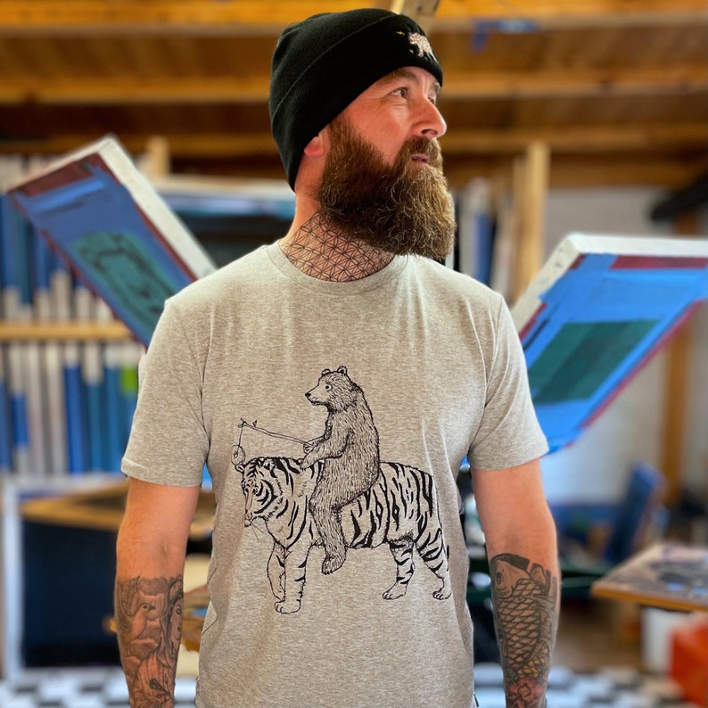 May include: A gray t-shirt with a black and white illustration of a bear riding a tiger. The bear is holding a stick with a string attached to it. The tiger has stripes and is looking to the left.