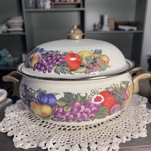 Puede incluir: Cazuela de esmalte color crema con tapa, con un diseño de frutas vibrantes de manzanas, uvas y peras. El plato tiene asas doradas y un pomo de tapa a juego. Se encuentra sobre un tapete de encaje blanco.