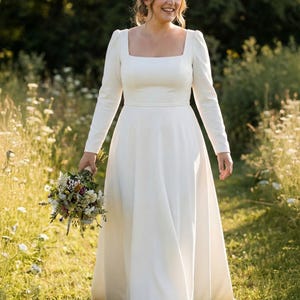 May include: A white wedding dress with a square neckline and long sleeves. The bride is holding a bouquet of flowers and walking through a field of wildflowers. The dress has a fitted bodice and a flowing skirt.