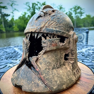 May include: A distressed, brown and bronze-toned helmet featuring a jagged, tooth-like aperture. The helmet rests on a wooden base, with a blurred natural backdrop of trees and water.