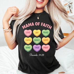 Puede incluir: Camiseta negra con la frase rosa "MAMA OF FAITH". Debajo, nueve caramelos en forma de corazón con palabras como "LOVE", "JOY" y "PEACE". El texto "Proverbios 31:28" está en la parte inferior. Una persona lleva la camiseta.