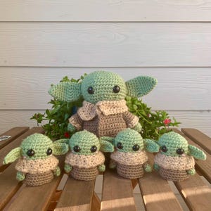 May include: A family of five crocheted figures, resembling a popular character. The figures are in varying sizes, with green heads, large ears, and black eyes. They wear tan and brown clothing and are arranged on a wooden surface.