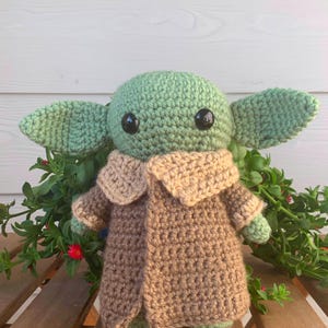May include: A handmade crochet doll in the likeness of a popular character. The doll has a green head and large ears, black eyes, and is wearing a brown coat with a tan collar. The doll is sitting on a wooden surface with green foliage.