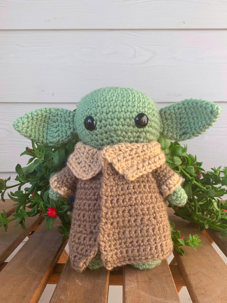 May include: A handmade crochet doll in the likeness of a popular character. The doll has a green head and large ears, black eyes, and is wearing a brown coat with a tan collar. The doll is sitting on a wooden surface with green foliage.
