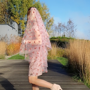 Peut inclure: Un voile rose clair orné d'un motif de petits cœurs rouges brodés, associé à une robe rose clair assortie et des bottines beiges. Le voile est confectionné en tulle transparent et tombe sur la tête et les épaules dans un style superposé.