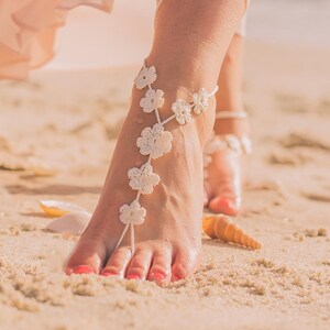 May include: White crocheted barefoot sandals with flower embellishments. The sandals are worn on a woman's feet on a sandy beach.