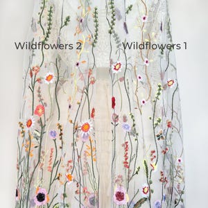May include: Sheer white fabric embroidered with colorful wildflowers. The design features various flowers in shades of pink, orange, yellow, and purple, along with green stems and leaves. The text "Wildflowers 2" and "Wildflowers 1" is visible.