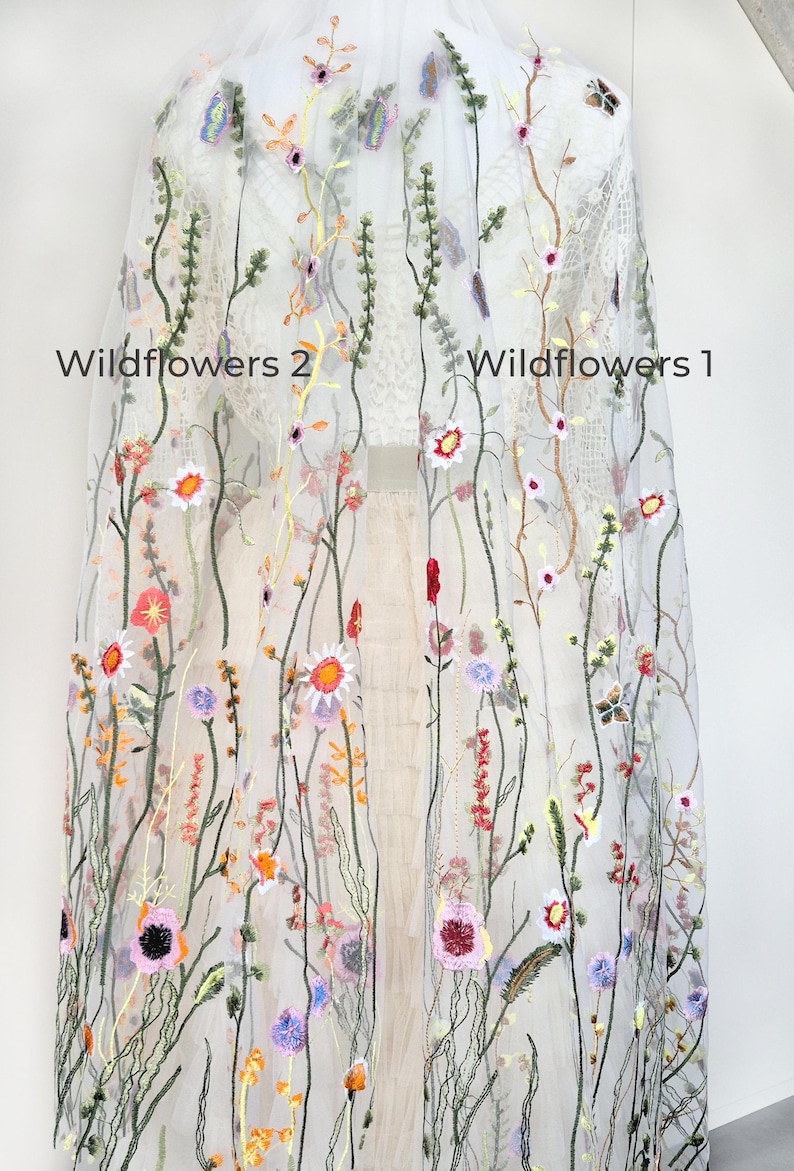 May include: Sheer white fabric embroidered with colorful wildflowers. The design features various flowers in shades of pink, orange, yellow, and purple, along with green stems and leaves. The text "Wildflowers 2" and "Wildflowers 1" is visible.