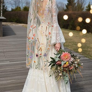 May include: A cream-colored wedding dress with floral embroidery on the veil and bodice. The dress features long lace sleeves and a layered skirt. A bouquet of flowers is held in the hand.