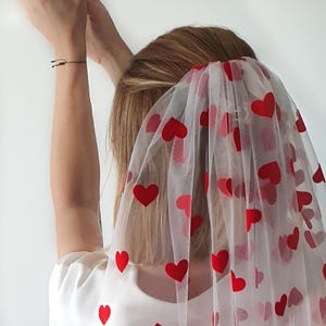 May include: A white tulle veil with red heart patterns. The veil is worn by a person with long blonde hair.