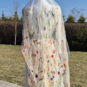 May include: A sheer, ivory-colored wedding veil with colorful floral embroidery. The veil features a delicate design of embroidered flowers in shades of red, orange, purple, and yellow, creating a whimsical and romantic look. The veil is long and flowing.