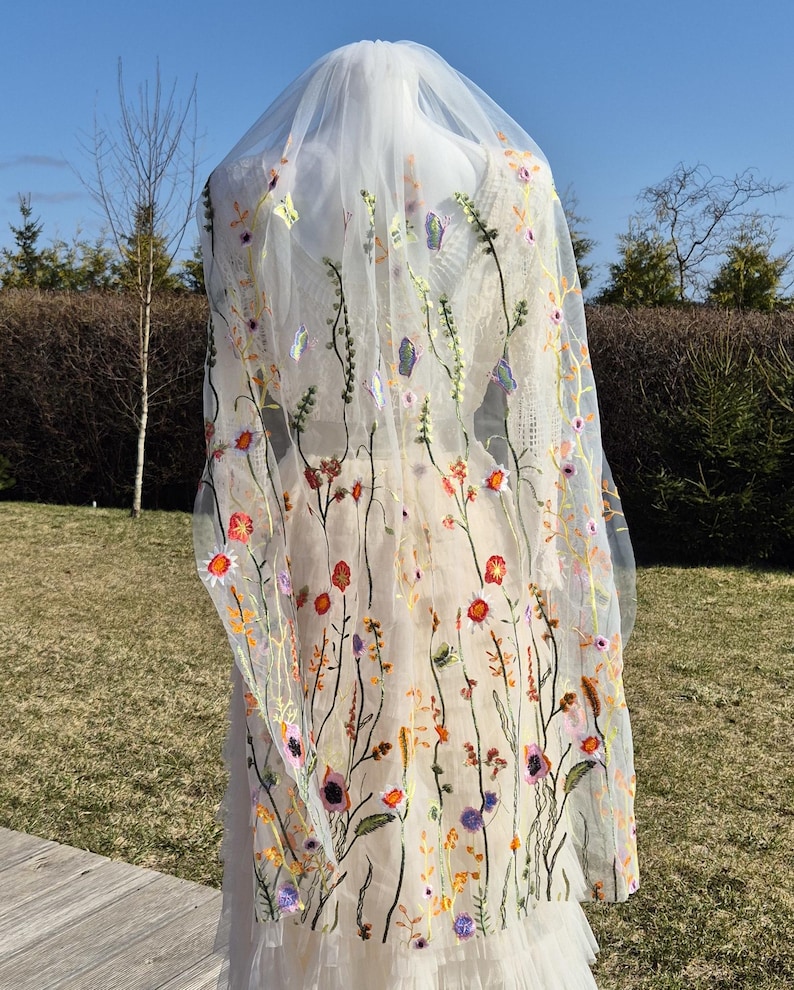 May include: A sheer, ivory-colored wedding veil with colorful floral embroidery. The veil features a delicate design of embroidered flowers in shades of red, orange, purple, and yellow, creating a whimsical and romantic look. The veil is long and flowing.