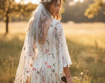 Puede incluir: Vestido de novia blanco con bordados florales y mangas de encaje. El vestido presenta un velo transparente con detalles florales a juego y una corona de flores de margarita. La novia sostiene un ramo de flores.