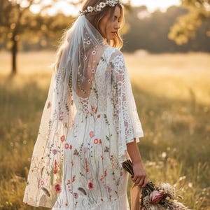 May include: A white lace wedding dress with floral embroidery and bell sleeves. The dress features a low back and a flowing skirt. A sheer veil with matching floral embroidery and a daisy crown complete the bridal look. A bouquet of dried flowers is held.