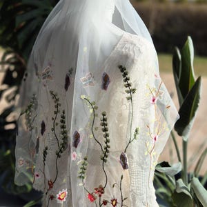 May include: A white wedding veil with delicate floral embroidery. The veil features embroidered flowers, butterflies, and vines in shades of red, pink, green, and purple. The veil is draped over a white dress.