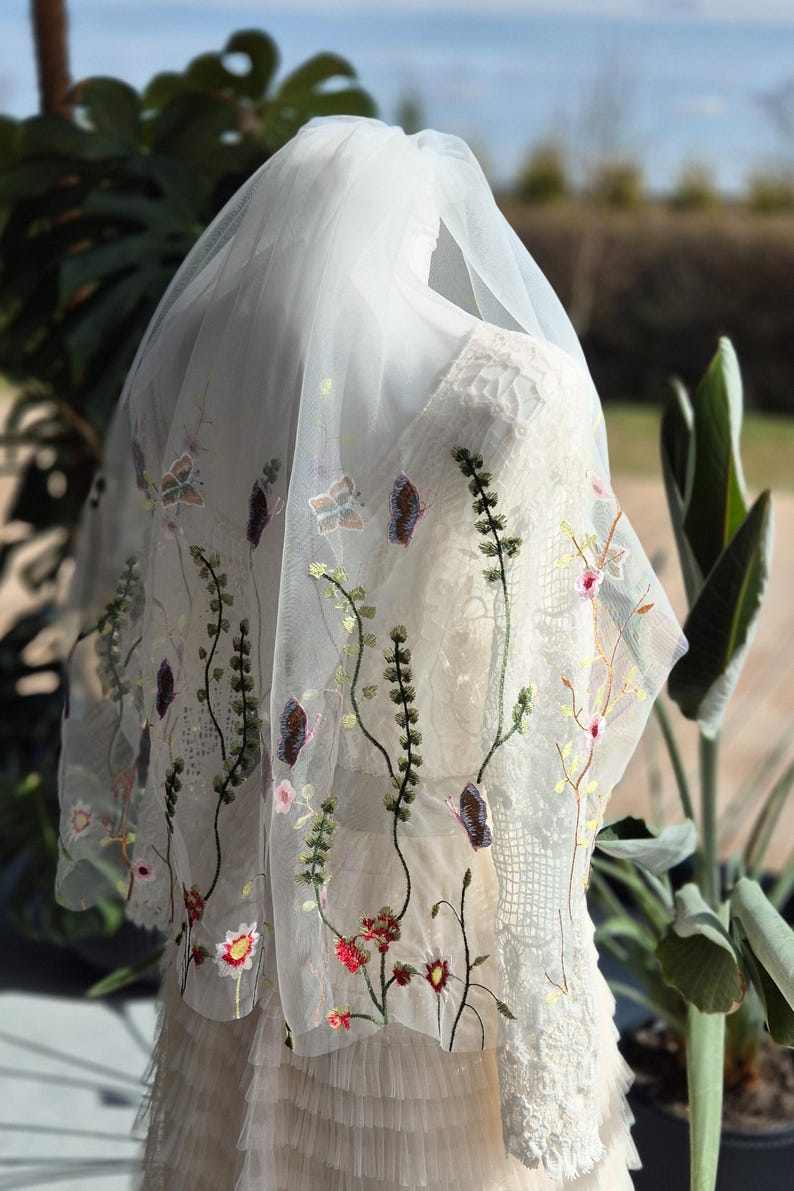 May include: A white wedding veil with delicate floral embroidery. The veil features embroidered flowers, butterflies, and vines in shades of red, pink, green, and purple. The veil is draped over a white dress.