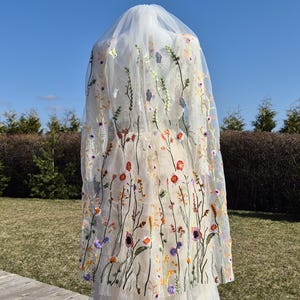 May include: A sheer white wedding veil with colorful embroidered flowers and butterflies. The veil is long and flowing, with a delicate floral design in shades of red, orange, purple, and yellow.