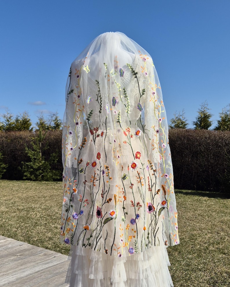 May include: A sheer white wedding veil with colorful embroidered flowers and butterflies. The veil is long and flowing, with a delicate floral design in shades of red, orange, purple, and yellow.