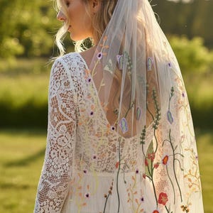 May include: A white wedding dress with a floral embroidered veil. The dress features a lace bodice and long sleeves. The veil is adorned with colorful embroidered flowers and greenery, creating a romantic and whimsical look.