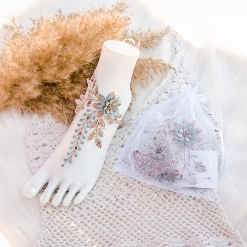 May include: A pair of delicate, floral ankle jewelry with a light blue and brown design. The jewelry is made of lace and features a floral pattern with small beads. The jewelry is displayed on a white mannequin foot.