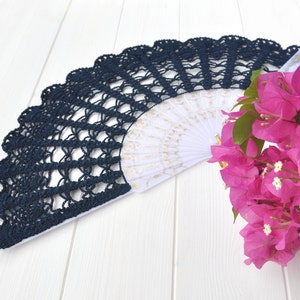 May include: A navy blue crocheted fan with a white and gold patterned interior. The fan is lying on a white wooden surface next to a bunch of pink bougainvillea flowers.