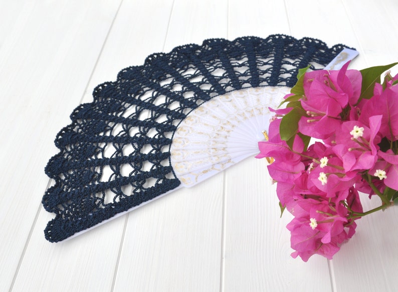 May include: A navy blue crocheted fan with a white and gold patterned interior. The fan is lying on a white wooden surface next to a bunch of pink bougainvillea flowers.