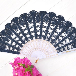 May include: A navy blue crocheted fan with a white and gold fan inside. The fan is on a white wooden surface.