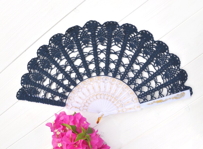 May include: A navy blue crocheted fan with a white and gold fan inside. The fan is on a white wooden surface.