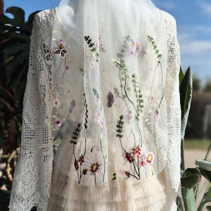 May include: Ivory wedding dress with a layered skirt and lace sleeves. The dress is adorned with embroidered flowers and butterflies on the sheer overlay. The veil is draped over the shoulders, creating a romantic and ethereal look.
