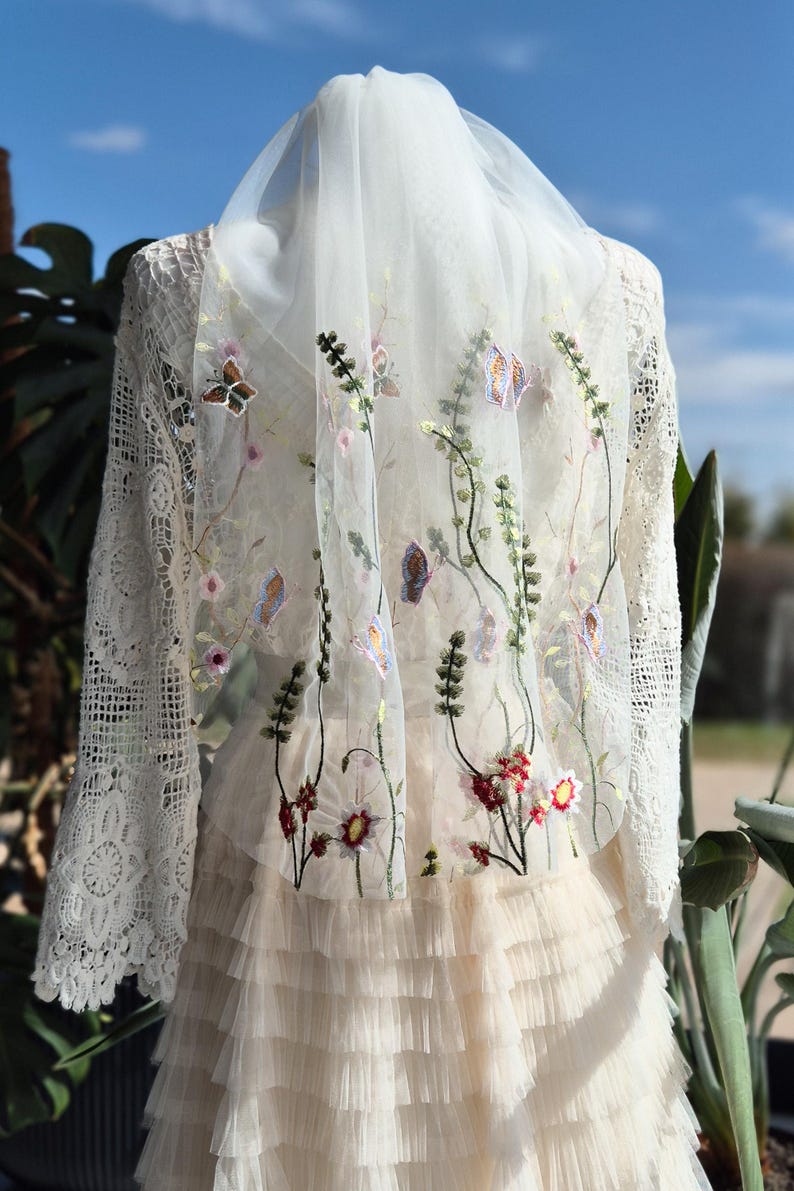 May include: Ivory wedding dress with a layered skirt and lace sleeves. The dress is adorned with embroidered flowers and butterflies on the sheer overlay. The veil is draped over the shoulders, creating a romantic and ethereal look.