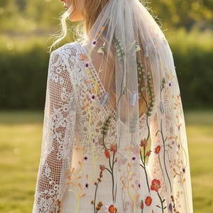 May include: A bride wearing a white wedding dress with a floral embroidered veil. The veil and dress feature colorful embroidered flowers in shades of orange, purple, and yellow. The dress has a lace bodice and long sleeves.