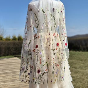 May include: A white wedding dress with a tulle skirt and a sheer floral embroidered cape. The cape features colorful embroidered flowers and greenery. The dress is displayed on a mannequin outdoors against a blue sky.