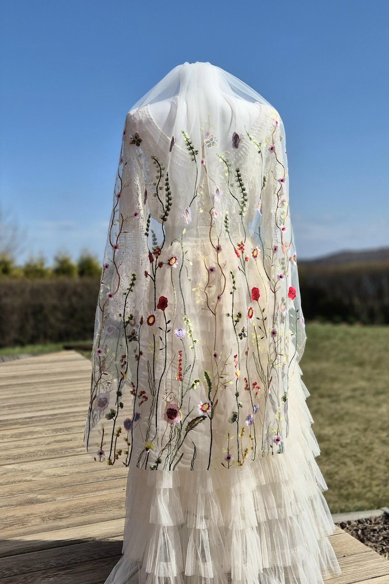 May include: A white wedding dress with a tulle skirt and a sheer floral embroidered cape. The cape features colorful embroidered flowers and greenery. The dress is displayed on a mannequin outdoors against a blue sky.
