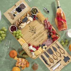 May include: A bamboo charcuterie board with a variety of cheeses, crackers, fruits, and meats. The board is engraved with "MR. & MRS. WILLIAMS EST. 2025". A bottle of rose champagne and a set of cheese knives are also present.