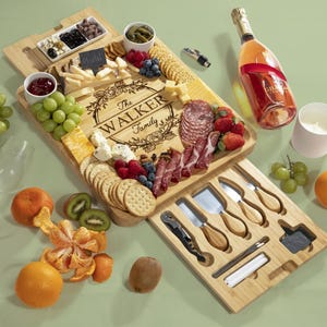 May include: A bamboo charcuterie board with the engraved text "The Walker Family" is filled with cheese, crackers, fruit, and meats. A bottle of champagne and a white candle are also present. A set of cheese knives is included.
