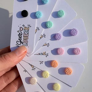 Könnte beinhalten: Eine Sammlung farbenfroher Ohrstecker in verschiedenen Pastelltönen, darunter Schwarz, Blau, Grün, Lila, Rosa, Orange und Gelb. Jedes Paar ist auf einer weißen Karte befestigt. Die Karten werden zusammengehalten und zeigen die Ohrringe.