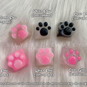 Puede incluir: Una colección de teclas con forma de pata, semiopacas y translúcidas, en varios colores, incluyendo rosa, blanco, beige y gris. Las teclas tienen un diseño de huella de pata y están etiquetadas. El texto "SILVERSCREAMKEYS" también es visible.