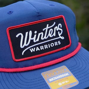 May include: Blue trucker hat with a red rope detail and a rectangular patch. The patch is black with the white text "Winters Warriors" and a red border. A small sticker is visible on the hat.