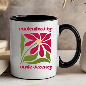 May include: White ceramic mug with a black interior and handle. The mug features a red flower design with green leaves and the text "radicalized by basic decency" in pink. The mug is displayed on a light-colored surface.