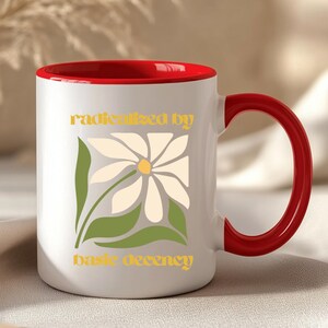 May include: White ceramic mug with a red interior and handle. The mug features a daisy design with the words "radicalized by basic decency" in yellow lettering. The mug is on a neutral surface.