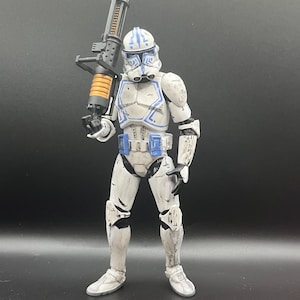 Pu&ograve; includere: Una action figure bianca e blu di un soldato che tiene in mano una grande arma nera e arancione. La figura ha un elmetto con segni blu e indossa un'armatura bianca con accenti blu. La figura &egrave; in piedi su una superficie scura.