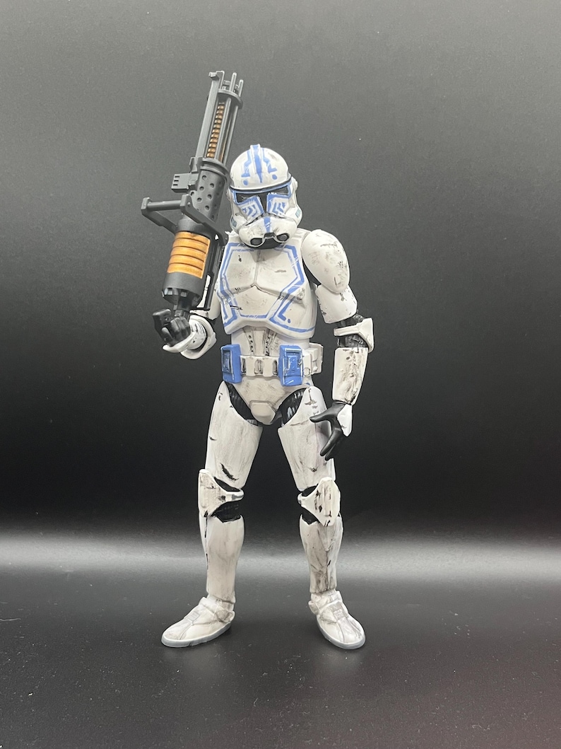 Pu&ograve; includere: Una action figure bianca e blu di un soldato che tiene in mano una grande arma nera e arancione. La figura ha un elmetto con segni blu e indossa un'armatura bianca con accenti blu. La figura &egrave; in piedi su una superficie scura.