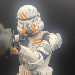 Pu&ograve; includere: Action figure dettagliata di un soldato corazzato bianco e arancione. L'elmo e l'armatura presentano strisce e segni arancioni, con un aspetto usurato. La figura tiene in mano un'arma, con sfondo nero.