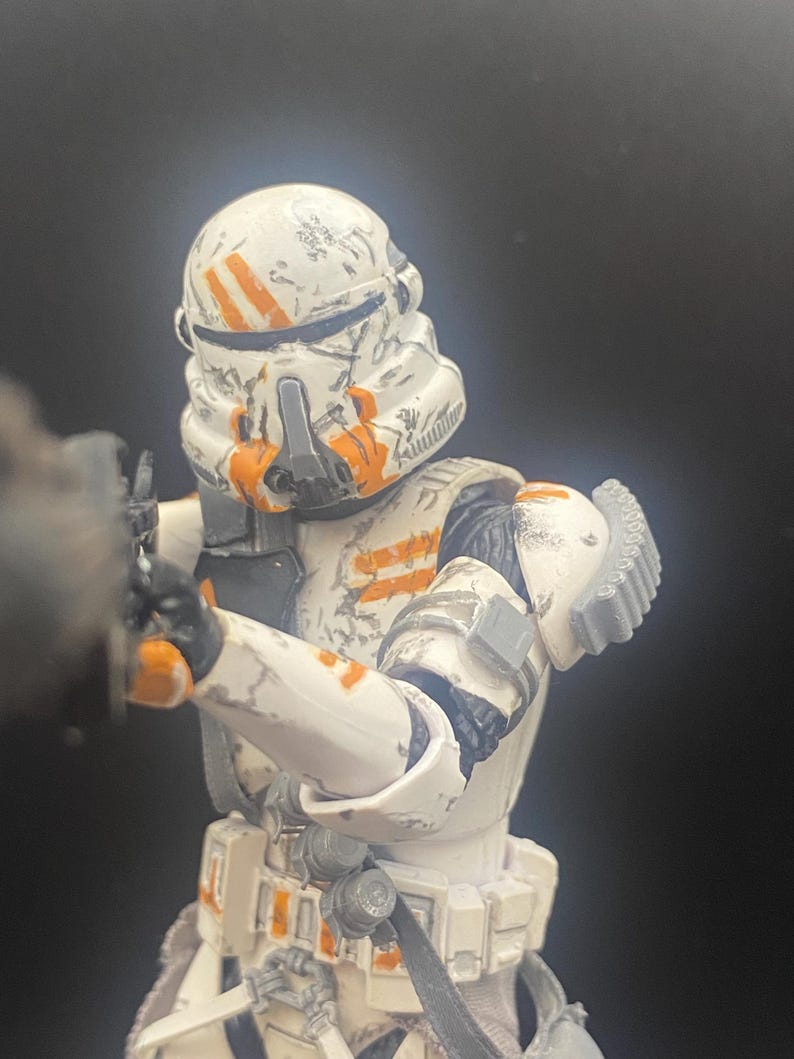 Pu&ograve; includere: Action figure dettagliata di un soldato corazzato bianco e arancione. L'elmo e l'armatura presentano strisce e segni arancioni, con un aspetto usurato. La figura tiene in mano un'arma, con sfondo nero.