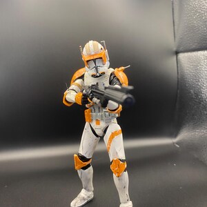 Pu&ograve; includere: Una action figure bianca e arancione di un clone trooper, che tiene in mano un fucile blaster nero. La figura ha un elmetto con segni arancioni e dettagli dell'armatura. La figura &egrave; in posizione di combattimento.